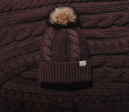 A brown woven beanie with a pom pom on top of the same brown woven fabric. 