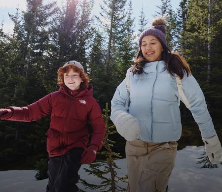 A boy in a red jacket and a girl in a light blue jacket running with pine trees behind them. 