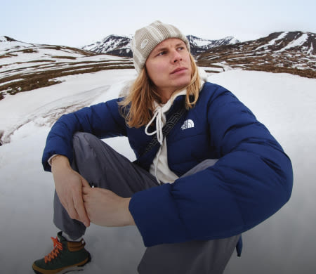 A guy in a white hoodie/tan beanie/navy jacket sitting in the snow. He’s looking away from the camera. 