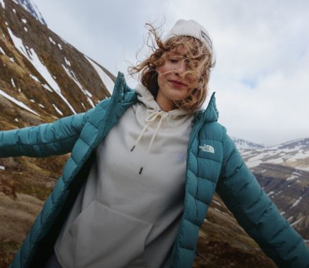 A woman in a white hoodie/white beanie/turquoise jacket with her arms stretched out.