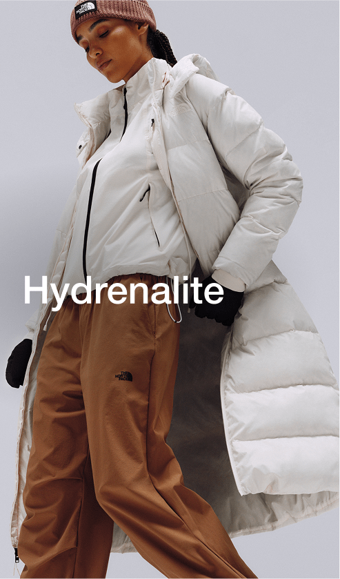 A female wearing The North Face Hydrenalite City Long Down Hooded Parka in White Dune and a brown knit beanie looks down against a neutral background. 