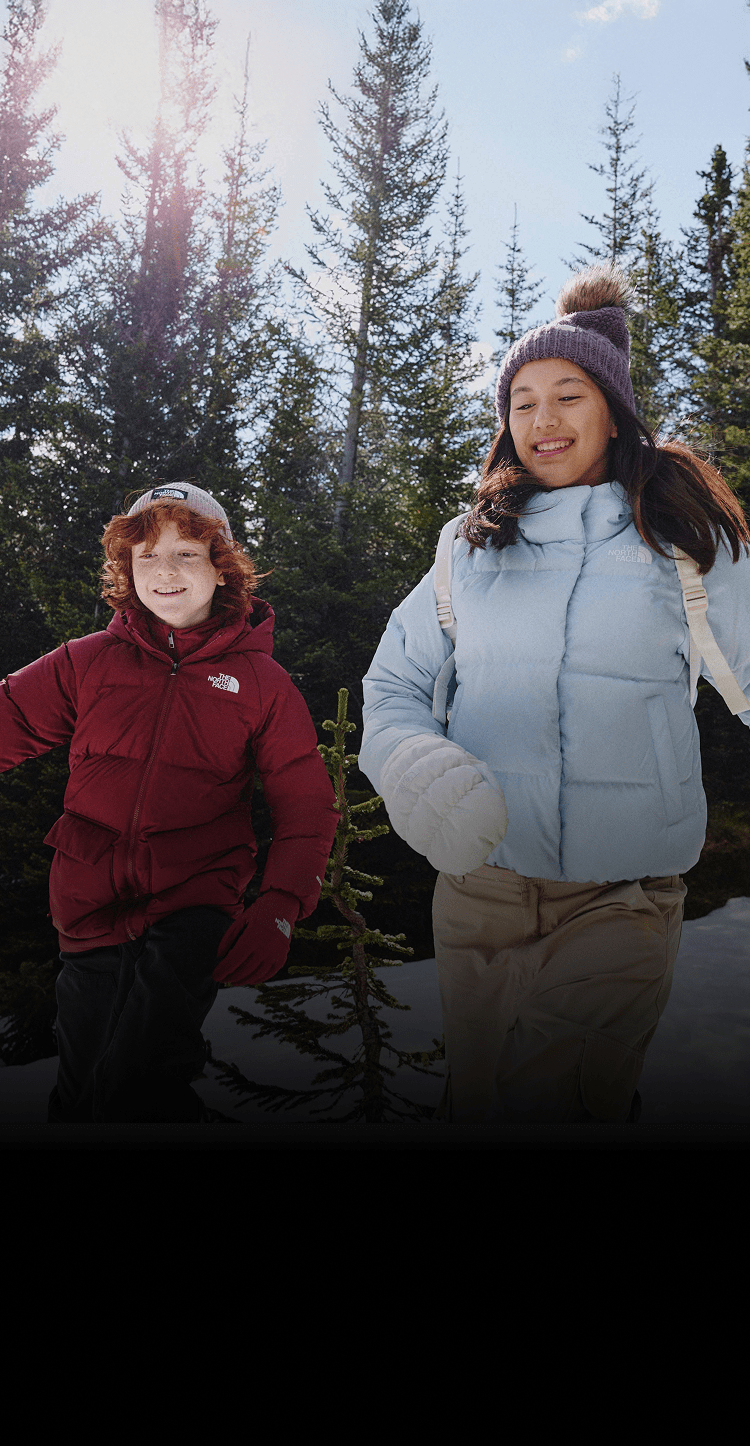 A boy in a red jacket and a girl in a light blue jacket running with pine trees behind them. 