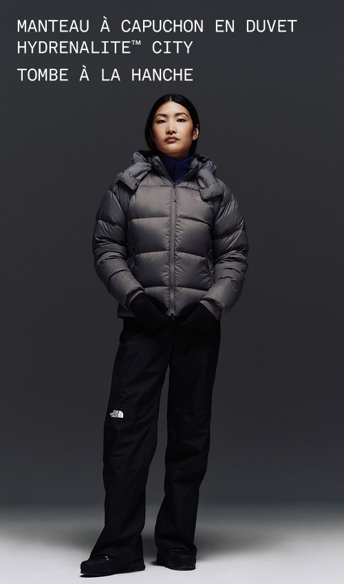 A female wearing The North Face Hydrenalite City Long Down Hooded Parka in White Dune and a brown knit beanie looks down against a neutral background. 