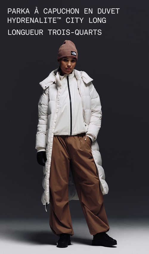 A female wearing The North Face Hydrenalite City Long Down Hooded Parka in White Dune and a brown knit beanie looks down against a neutral background. 