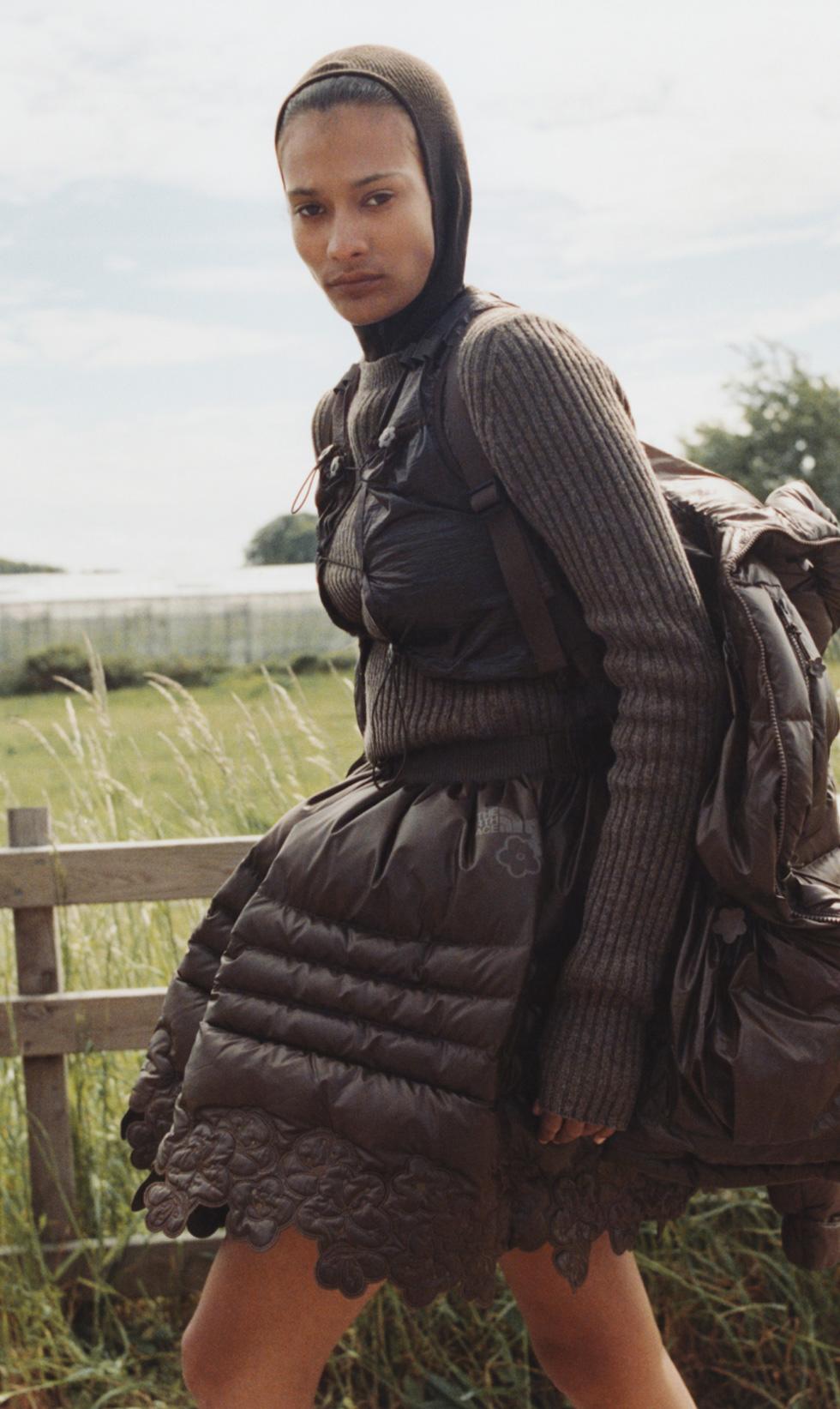 A woman in a brown knit top, brown skirt and brown backpack walking through a grassy field.
