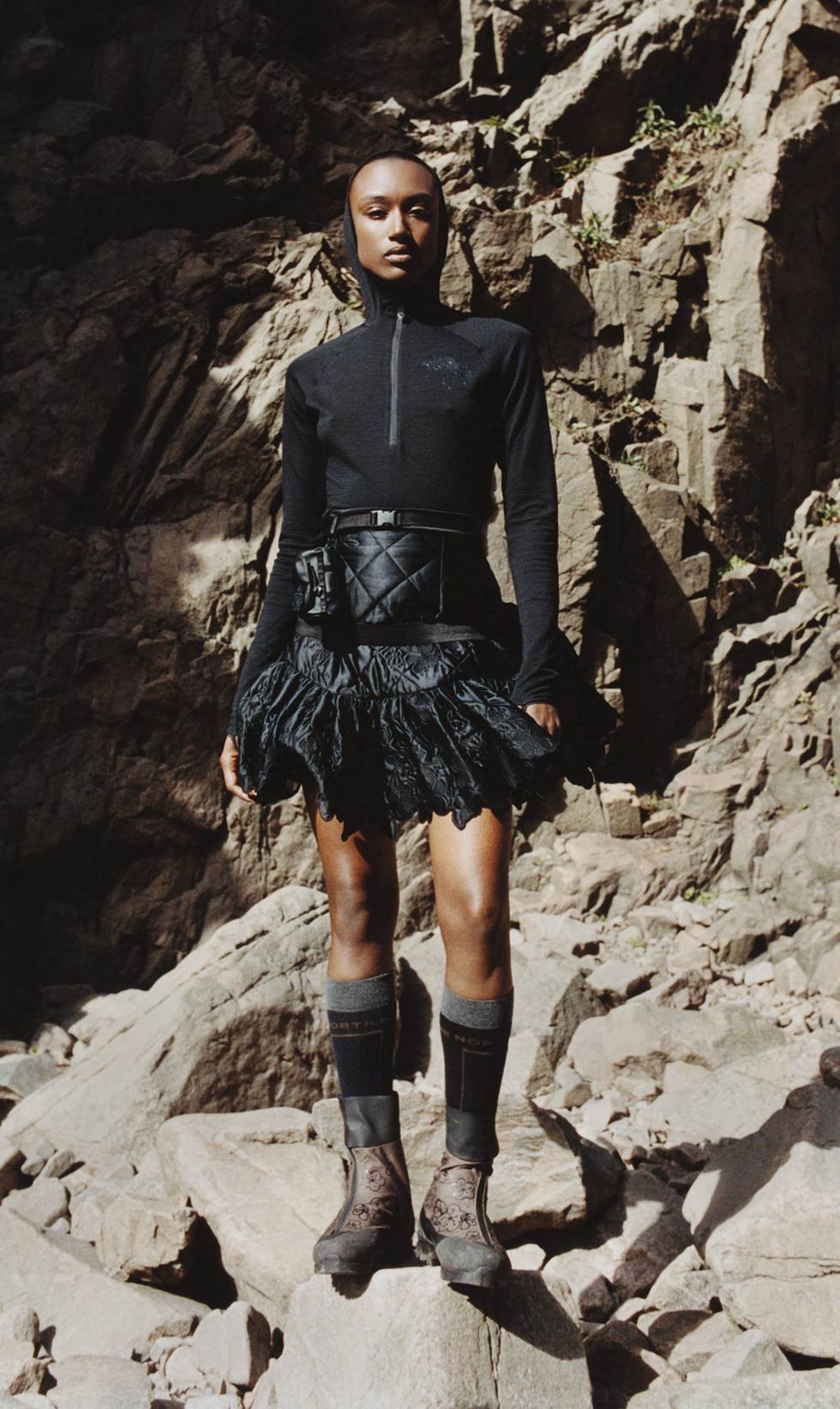 A woman in a black, half zip jacket and black skirt standing on some rocks looking at the camera.