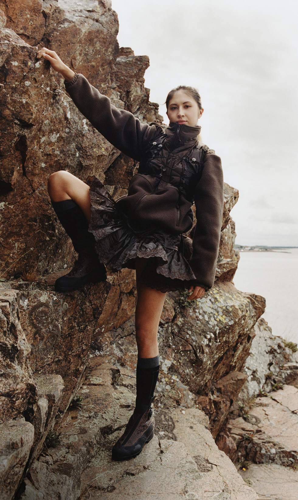 A woman in a brown fleece and skirt hanging off the side of a rock and looking at the camera. 