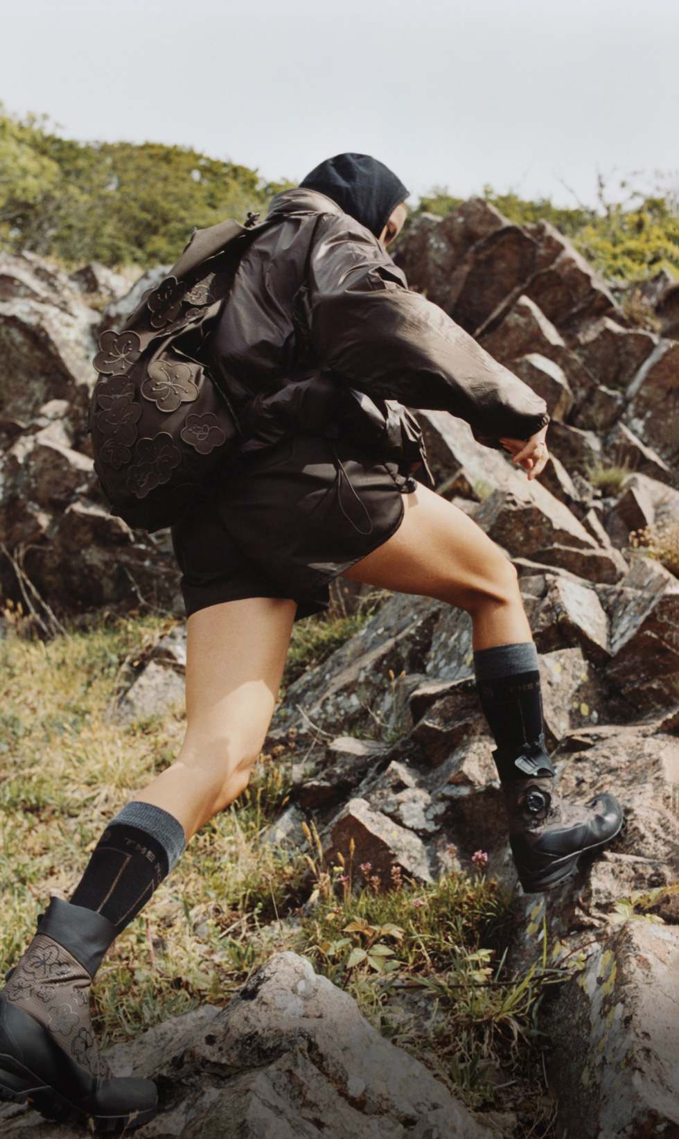 A woman with a brown, flowery backpack and a black hood up striding up some rocks on a grassy hill.