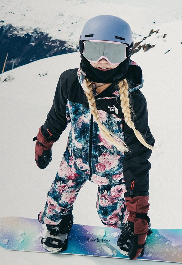 wo kids snowboard down a sunlit mountain in The North Face snow gear. A close-up on the right shows the girl smiling in her helmet and goggles, board in hand, ready to ride again. 