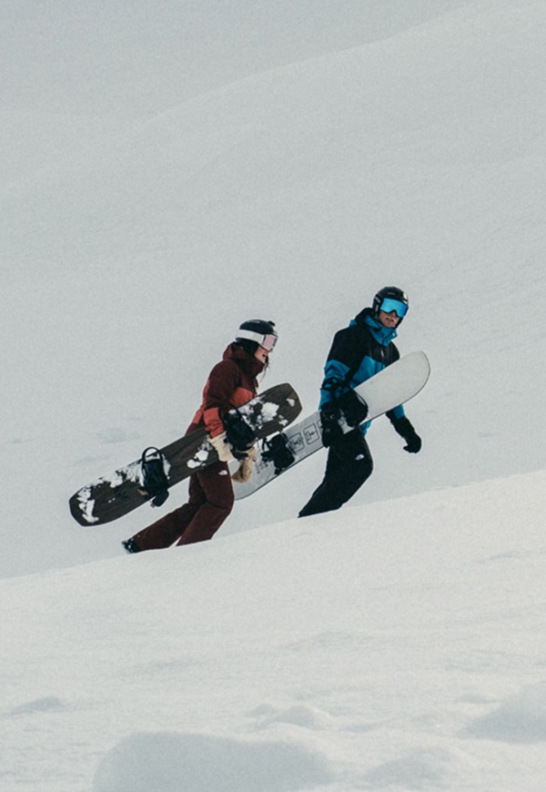Two snowboarders ride down a snowy mountain, the woman wearing The North Face Women’s THERMOBALL™ Snow Triclimate® Jacket in Sumac/Mars Dust. A third image shows them hiking uphill with their boards in hand, surrounded by fresh powder and mountain views. 