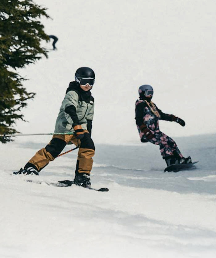 A guy in a white hoodie/tan beanie/navy jacket sitting in the snow. He’s looking away from the camera. 