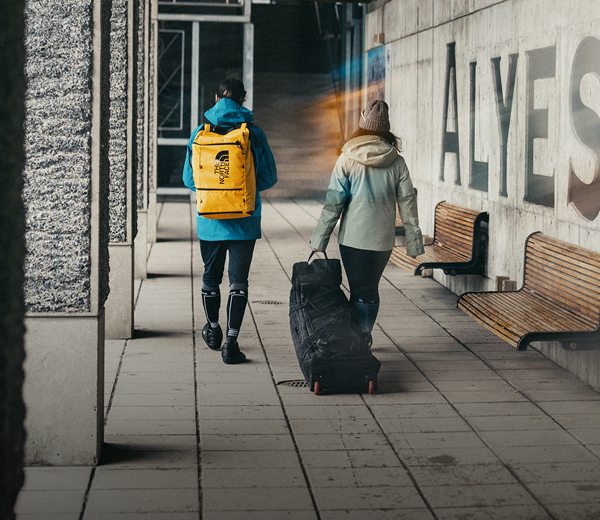 Deux skieurs déballent leur équipement du sac roulant Base Camp Snow Roller et du sac pour bottes Base Camp Boot Pack, souriant alors qu’ils enfilent leur manteau et se préparent pour une journée sur les pistes. 