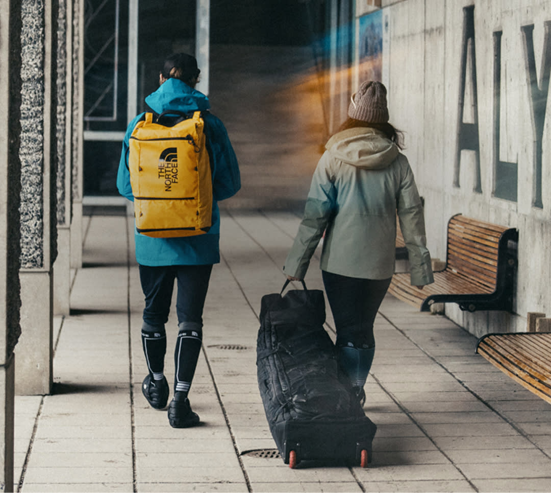 Two skiers unpack their gear from the Base Camp Snow Roller and Base Camp Boot Pack, smiling as they layer up and prepare for a day on the slopes.