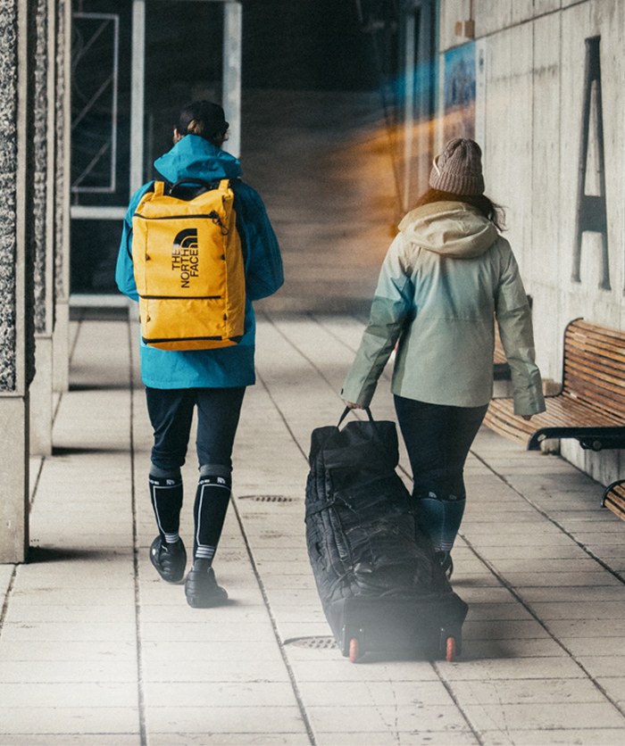 A boy in a red jacket and a girl in a light blue jacket running with pine trees behind them.