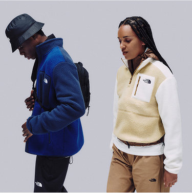 A man in a blue fleece and bucket hat with a cross-body bag facing left, and a woman white/beige fleece with black hair also facing slightly left.