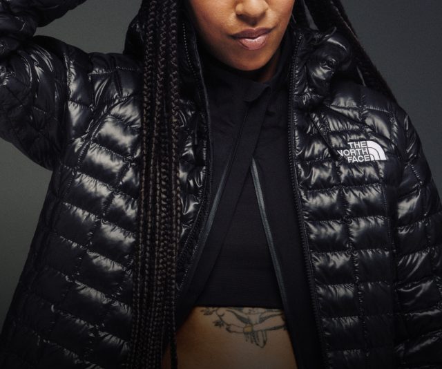 A man facing the camera in a black hat, cross-body bag and black puffer with a blue jacket underneath.