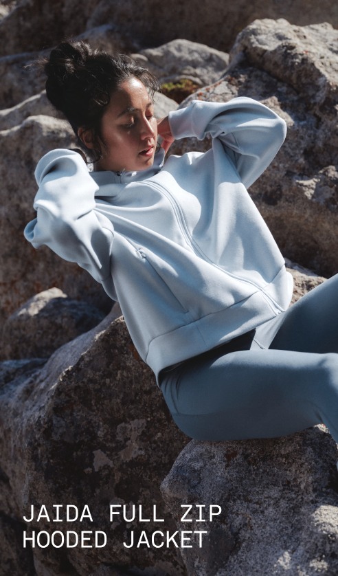 A model wearing the Jaida Full Zip Hooded Jacket in Granite Grey sits against a rock surface in an outdoor landscape.