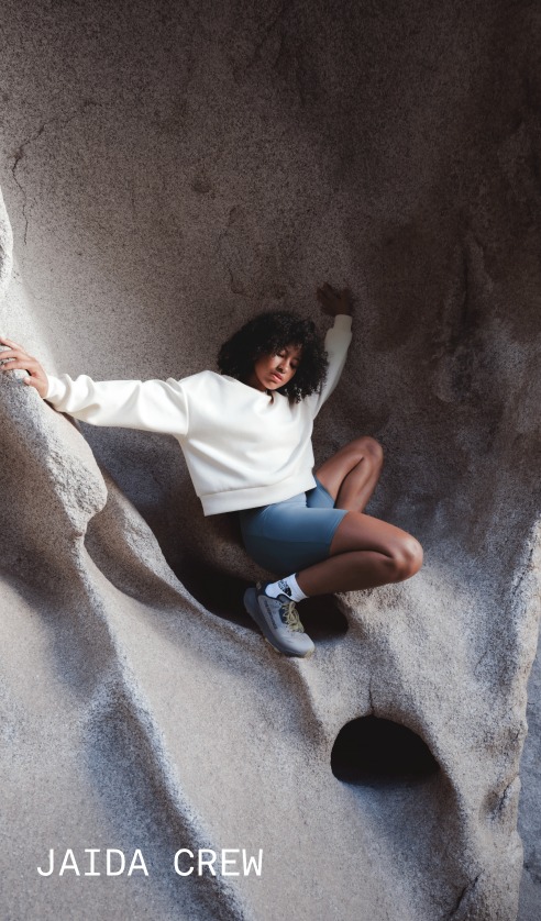 A model wearing the Jaida Crew in White Dune sits against a rock surface in an outdoor landscape.