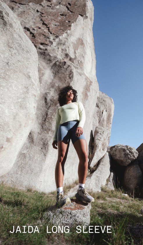 A model wearing the Jaida Long Sleeve in White Dune stands among large rock formations in an outdoor setting.