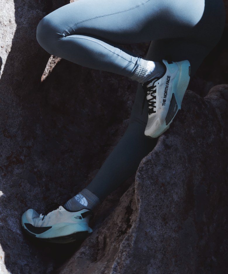 Close-up of an athlete’s legs wearing The North Face® Altamesa 500 sneakers in Opal Frost and Jaida Pocket Leggings in Granite Grey while stepping on rocks. 