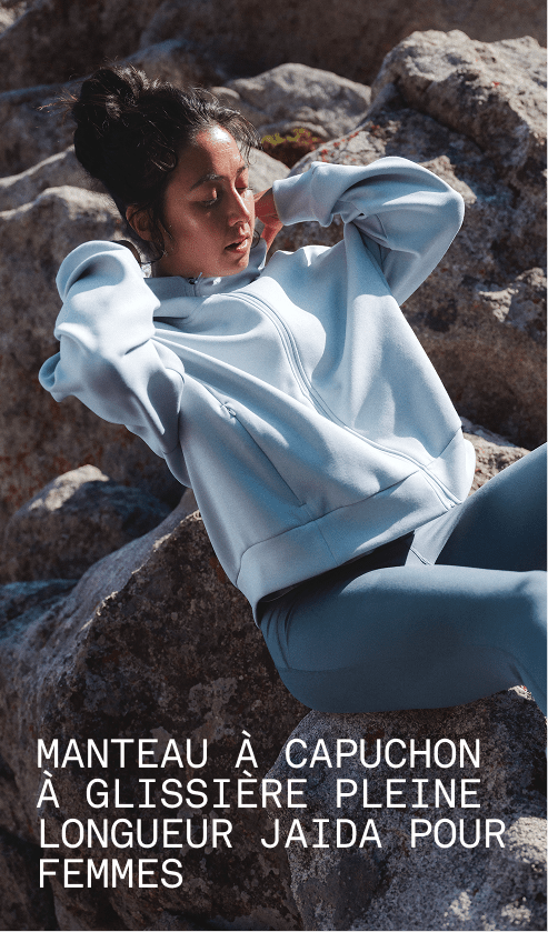 Un mannequin portant le manteau à capuchon Jaida à glissière pleine longueur en gris granite est assis contre une surface rocheuse dans un paysage extérieur.