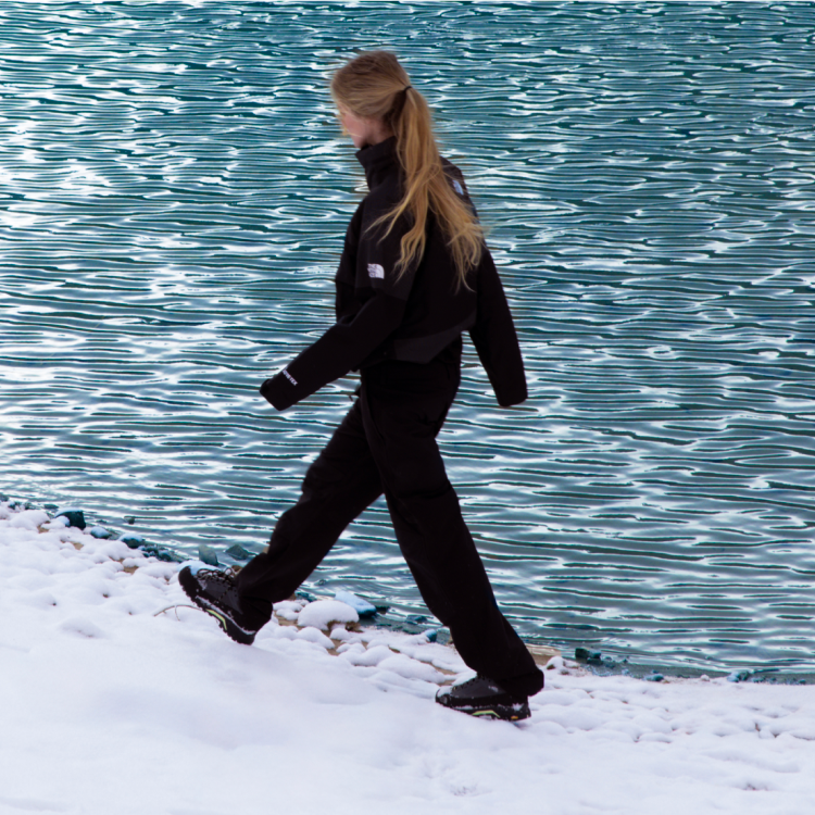 Woman in black jacket walks along lakeshore.