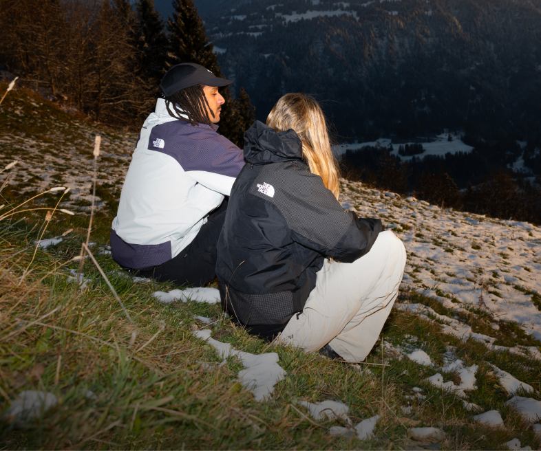 Man and woman in jackets sit on hillside.