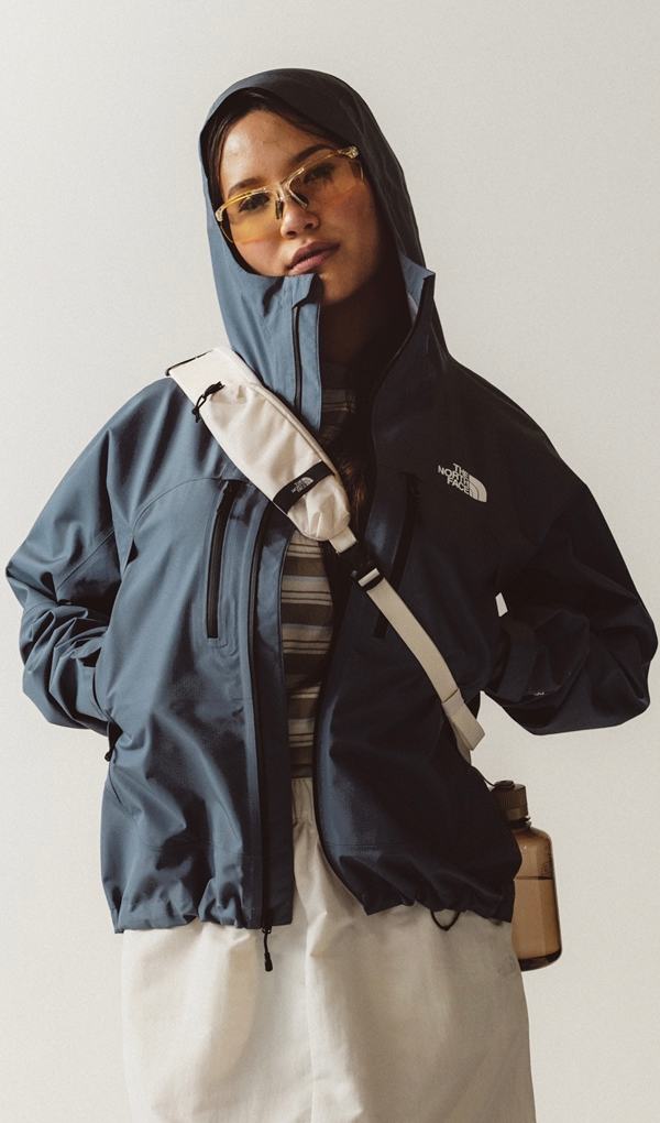 Women wearing yellow sunglasses leaning back in a blue rain jacket and white cross body pack. 