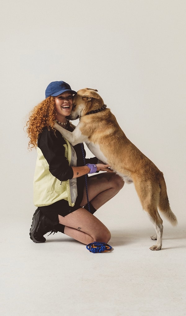 Woman playing with dog wearing white vest and blue hat. 