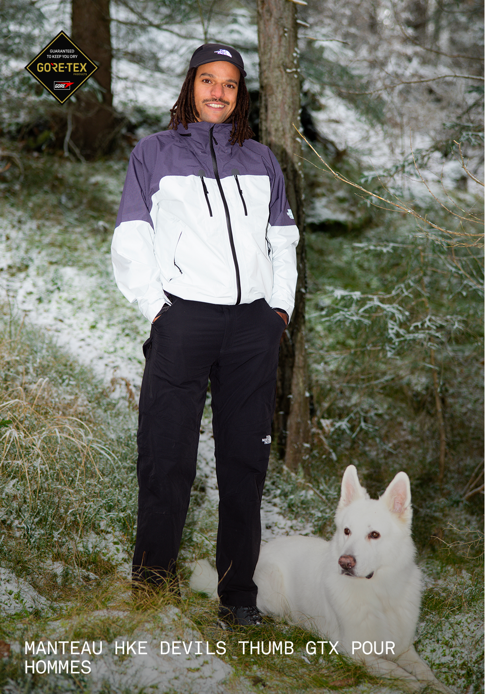 Homme en veste mauve et blanche debout près d’un chien.