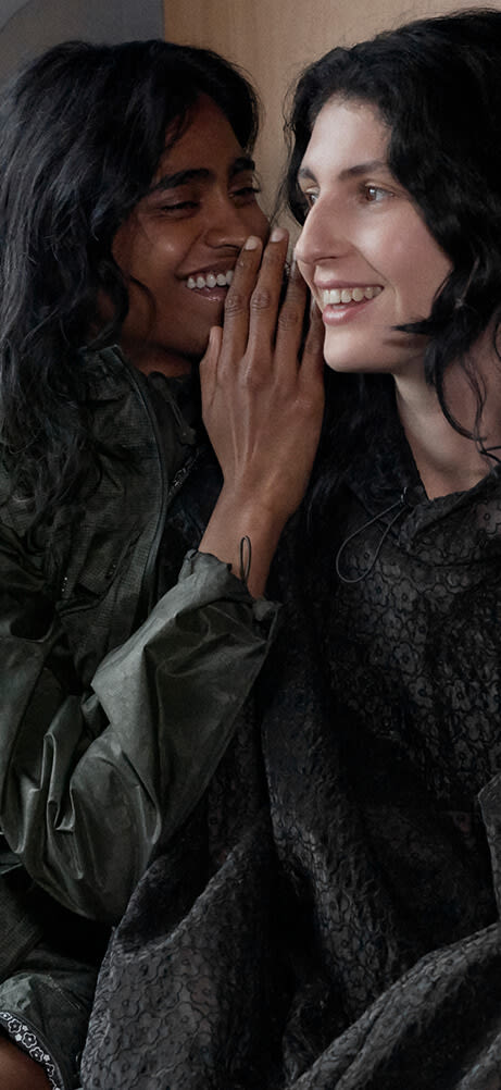 One model leans in to whisper as both laugh while sitting together on a train wearing The North Face x Cecilie Bahnsen collection with mountain scenery outside.