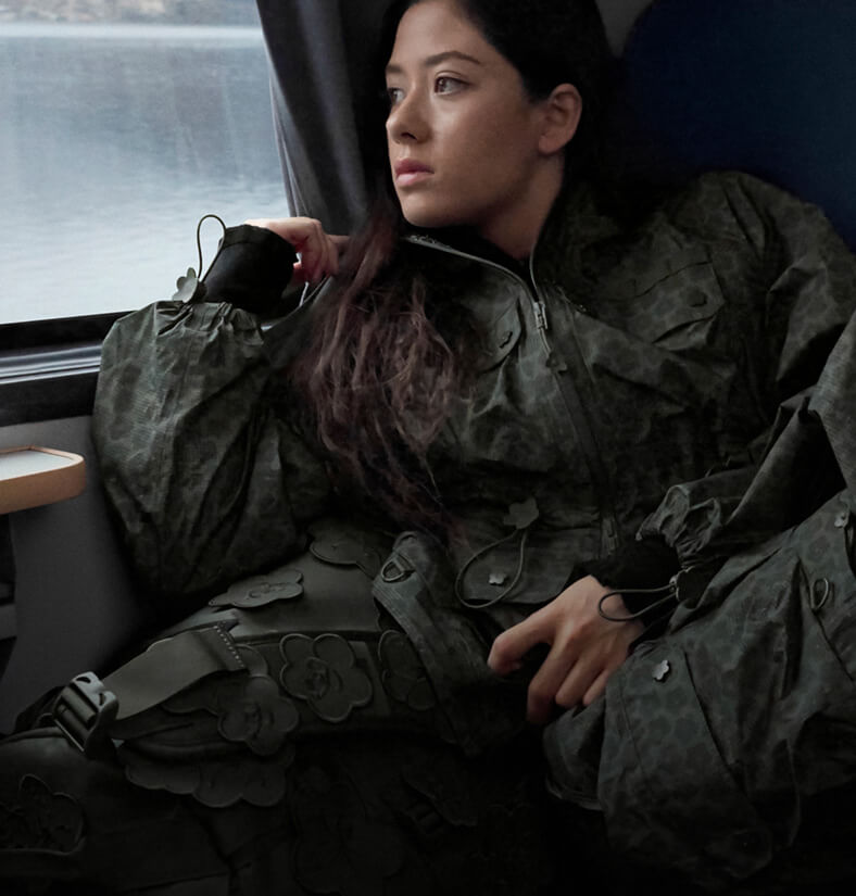 Model in The North Face x Cecilie Bahnsen collection relaxing on a train seat with alpine mountains visible through the window.