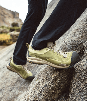 Closeup of a person’s legs, wearing black pants and yellow shoes. The person is on a steep rock face, hiking up.