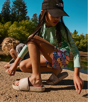 Child wearing a black hat, long-sleeved teal shirt, colorful shorts and beige slides, crouching down with a lake and woods in the background. 