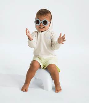 Baby wearing flower-shaped sunglasses, white hoodie and yellow shorts, sitting on a white stool, arms outstretched and facing the camera. 