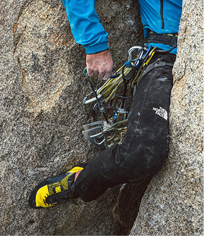 Side shot of a climber, face unseen, wedged between two rock faces. Person is wearing black pants and yellow climbing shoes with climbing gear attached to their harness. 