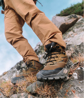 Person wearing brown hiking pants and brown hiking shoes with a black base. Closeup of the front of the shoes. Background is dry mountain terrain. 