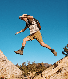 Man wearing an off-white shirt, brown shorts, and dark hiking shoes jumping between two rocks. 