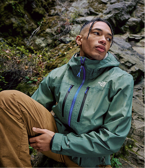 A person wearing a teal rain jacket and brown hiking pants, sitting on a rock. Rock wall with underbrush growing up the side. Climate is wet. 