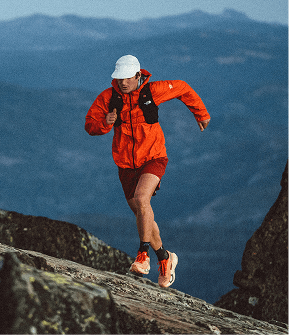 Man wearing an orange jacket, red shorts and white hat running up a rock slope. 