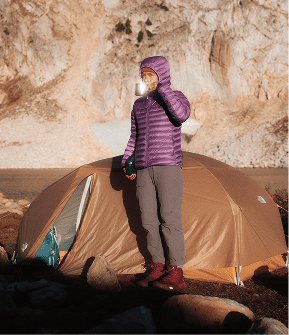 Woman wearing a purple hooded puffer jacket and gray pants standing in front of a brown tent drinking from a mug.