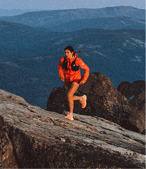 Woman standing on a mountain, facing away from the camera, wearing a teal jacket, teal shorts, white running shoes and black socks. 