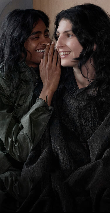 One model leans in to whisper as both laugh while sitting together on a train wearing The North Face x Cecilie Bahnsen collection with mountain scenery outside.