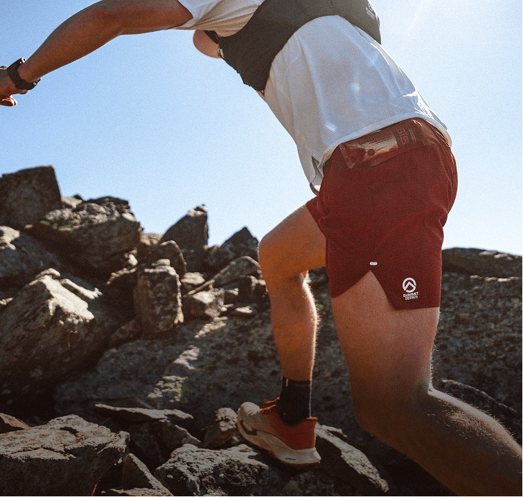 Side shot of person running up rocky terrain wearing a white shirt, black vest and red Pacesetter Shorts. 