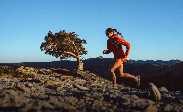 A woman running up a mountain in an orange jacket at sunset.