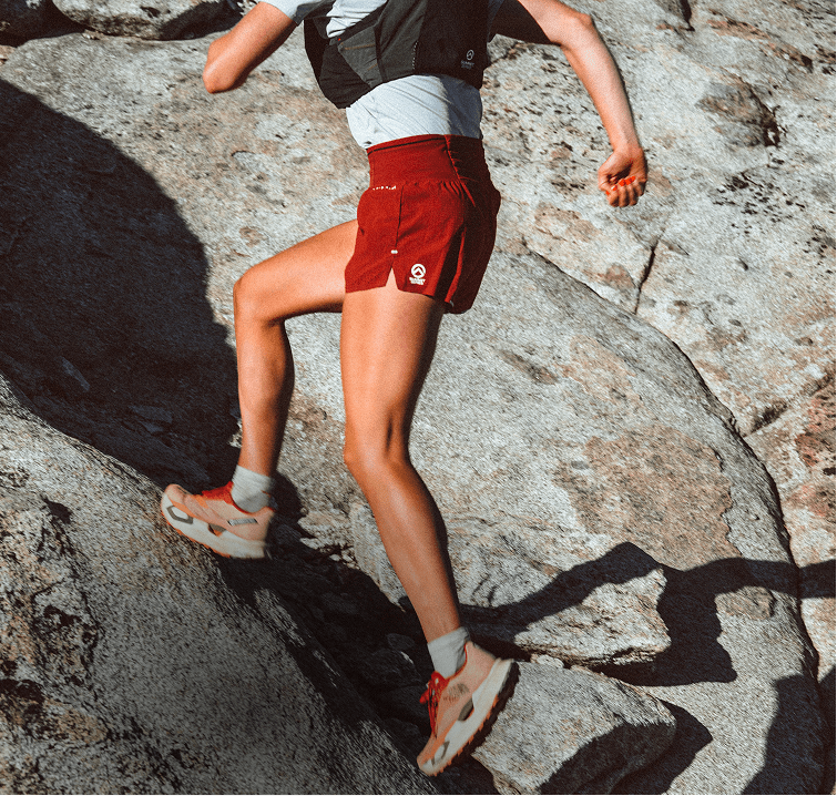 Side shot of person running up rocky terrain wearing a white shirt, black vest and red Pacesetter Shorts. 