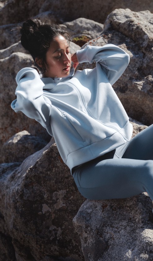 Athlete reclining on rocks wearing the Jaida Full Zip Hooded Jacket in Frost Grey and Jaida Pocket Leggings in Granite Grey in a rocky outdoor landscape. 
