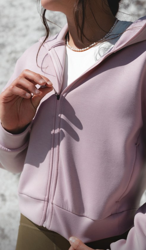 Athlete wearing Jaida Full Zip Hooded Jacket in a light pink tone, adjusting zipper outdoors near rock formations.