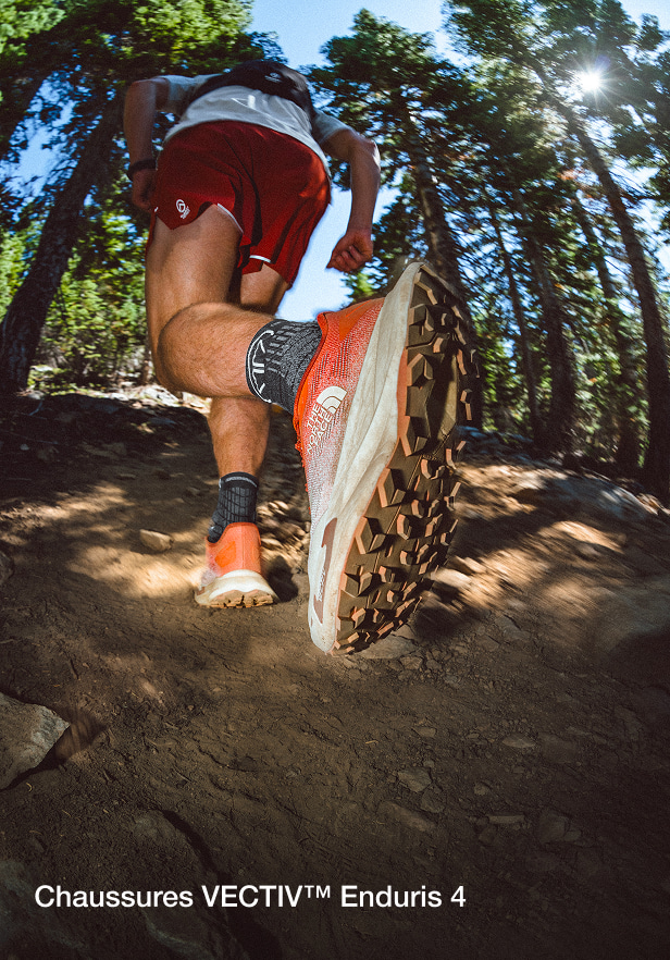 Gros plan sur l'arrière des chaussures Enduris 4, tandis qu'un homme court sur un sentier en forêt.