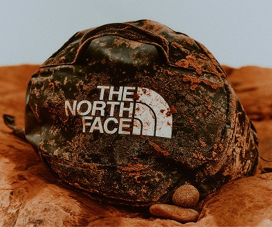 Side shot of a dusty black duffel bag laying on rocks with The North Face Logo. 